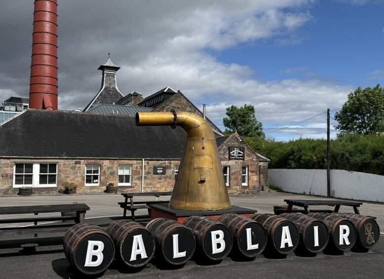 A large brass distilling apparatus with barrels labeled 'BALBLAIR' in front of a building with a red chimney. 5