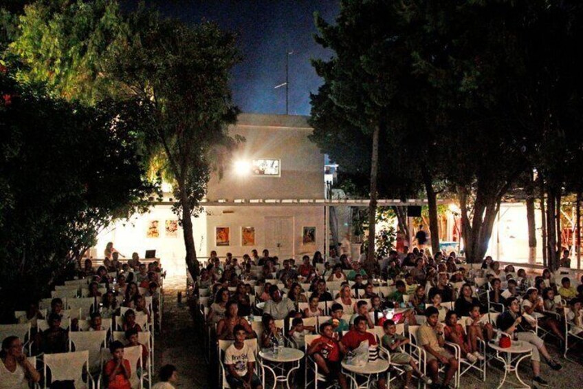 An outdoor gathering of people seated at round tables under trees, with a building in the background. 5