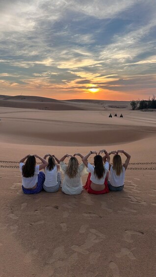 Vier Personen sitzen auf Sanddünen bei Sonnenuntergang, mit einer Wüstenlandschaft und einem Sonnenuntergang im Hintergrund. 1