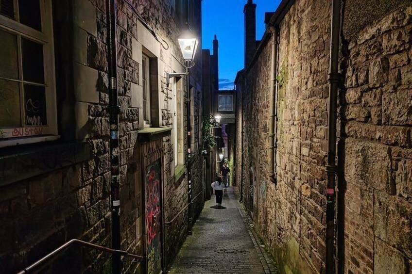 A narrow alleyway with stone walls, a person walking, and a street lamp. 2