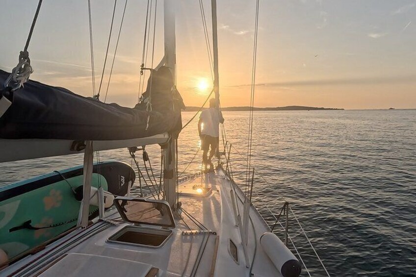 A sailboat with a person standing on the deck, a sunset in the background, and calm water. 5