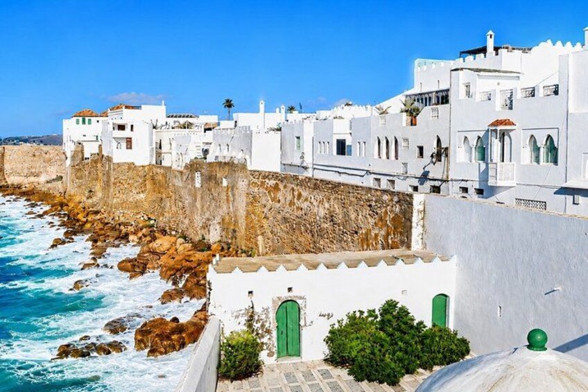 A coastal town with white buildings, a rocky shoreline, and a clear blue sky. 1