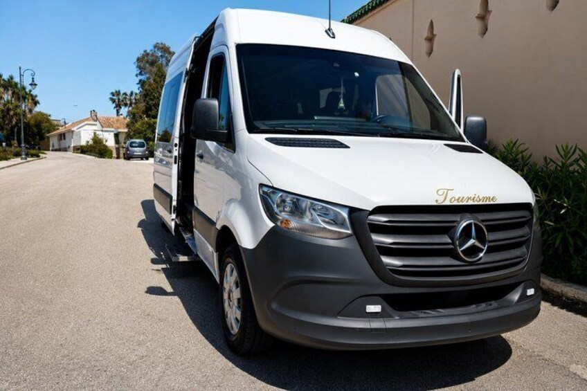 A white Mercedes van with 'Tourisme' on the front, parked on a street with houses and palm trees in the background. 5