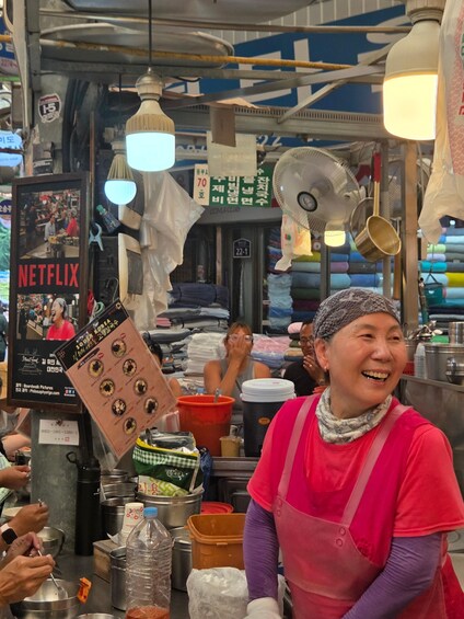 A person in a red apron at a market stall with various items like a fan, a poster, and stacked goods in the background. 4