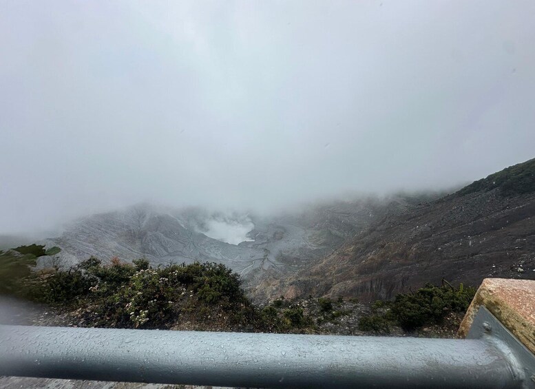 A mountainous landscape with a lake partially obscured by fog, a guardrail in the foreground, and sparse vegetation. 5