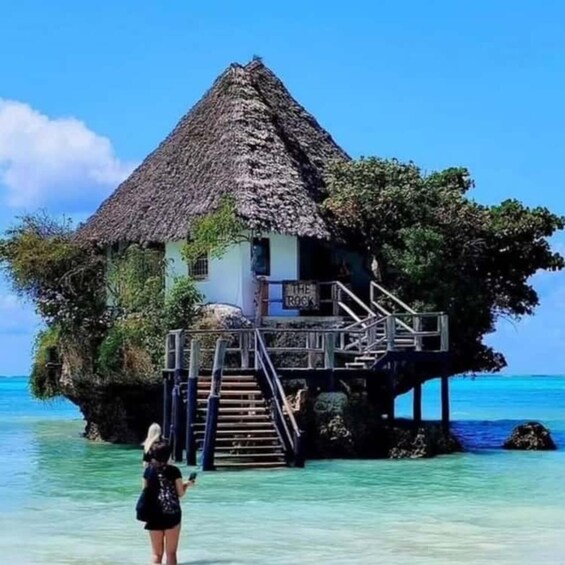 A thatched-roof house on stilts over clear turquoise water, with a sign reading 'The Rock.' 4