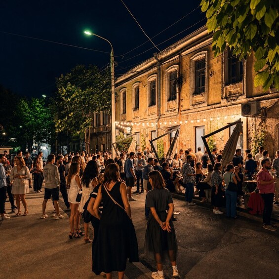 A crowded street at night with people gathered outside a building with string lights. 1