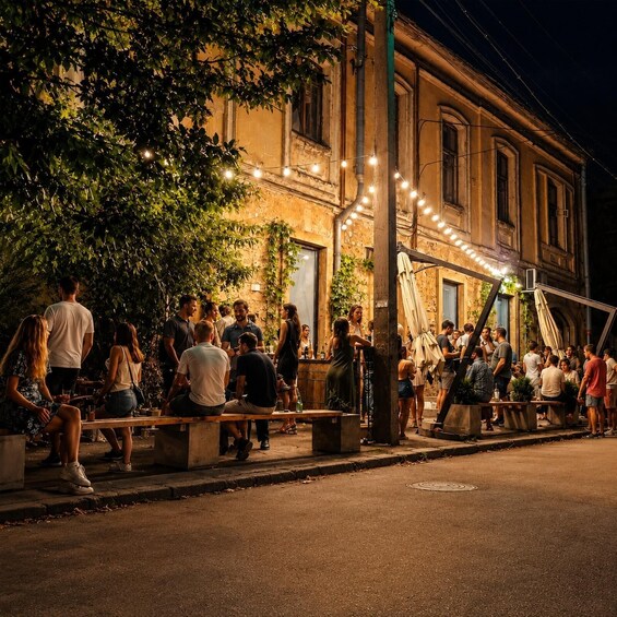 A crowded outdoor social gathering with people sitting and standing around tables and benches, under string lights, with a building in the background. 5