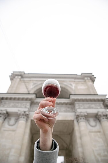 A hand holding a wine glass with a red liquid, in front of a large stone archway. 4