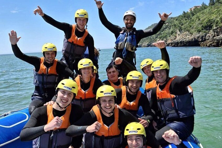A group of people on a blue raft wearing life jackets and helmets, with a rocky coastline in the background. 2
