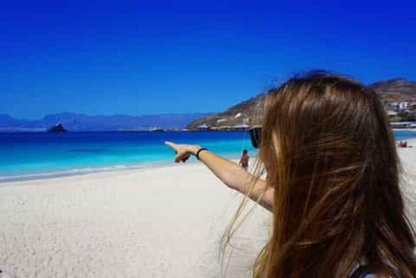 A beach with clear blue water, a sandy shore, and a person pointing towards something in the distance. 3