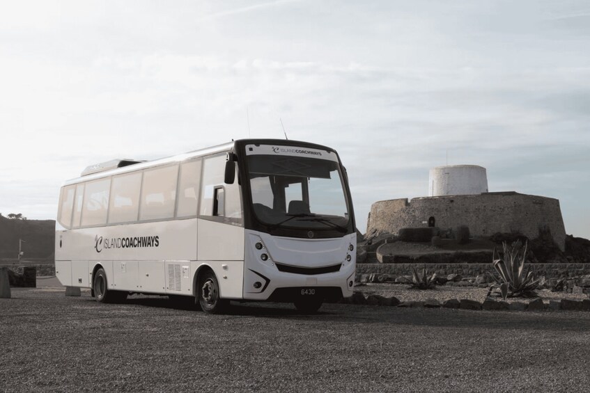 A white Island Coachways bus parked on a gravel surface near a historical stone structure. 1