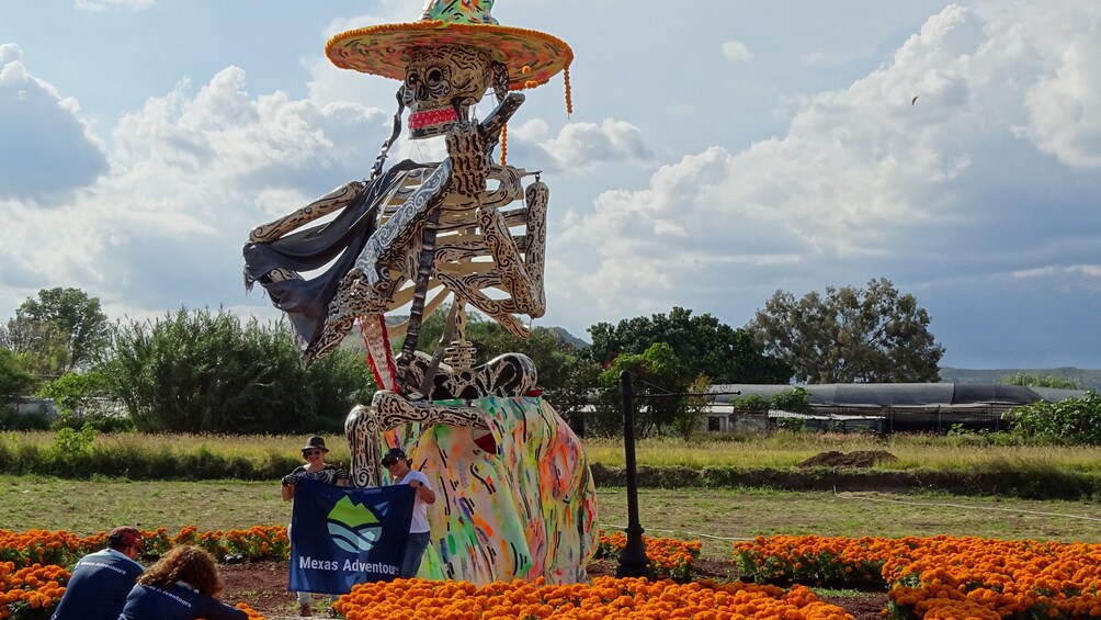 Una estatua colorida de un esqueleto con sombrero, rodeada de flores de cempasúchil. 5