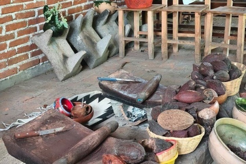 A rustic outdoor setting with various traditional tools and containers, including a large wooden mortar and pestle, a woven basket, and a clay pot. 4