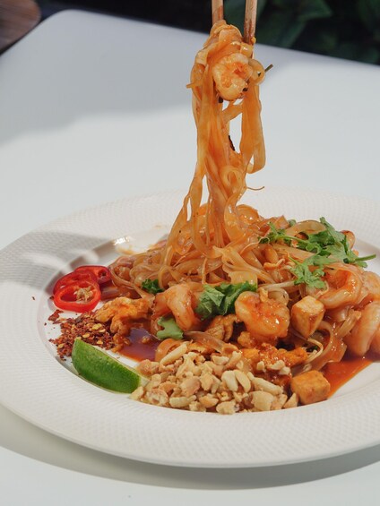 A plate of noodles with shrimp, garnished with cilantro and lime, and accompanied by sliced red chili and crushed peanuts. 3