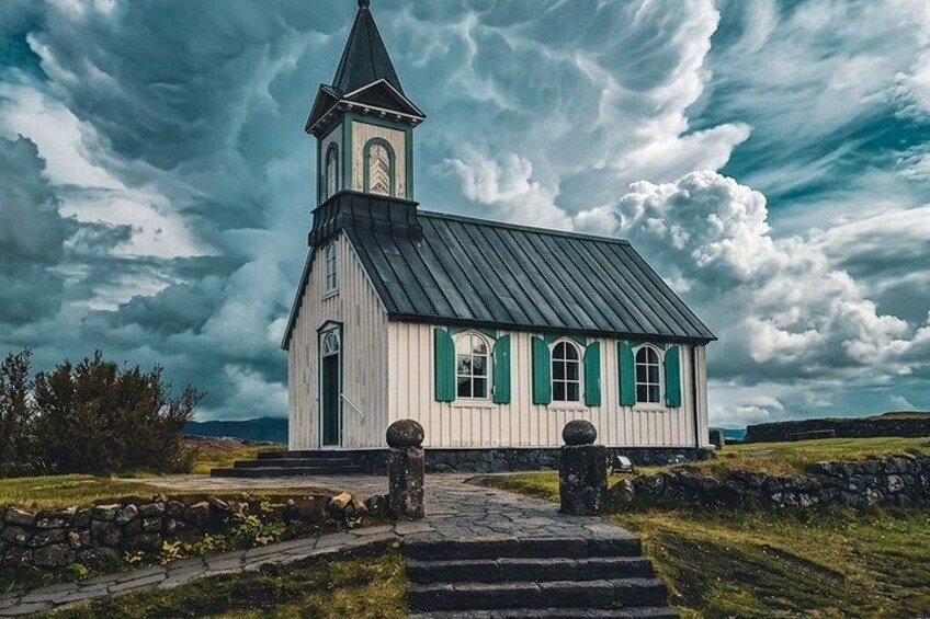 A white church with green shutters and a steeple, surrounded by stone walls and a stone pathway. 2