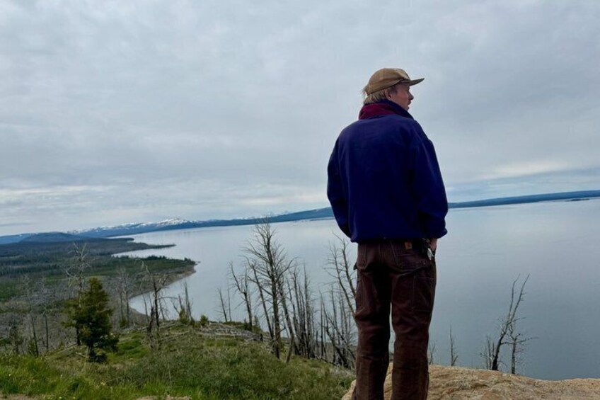 A person standing on a hill overlooking a calm lake with trees and distant mountains. 5