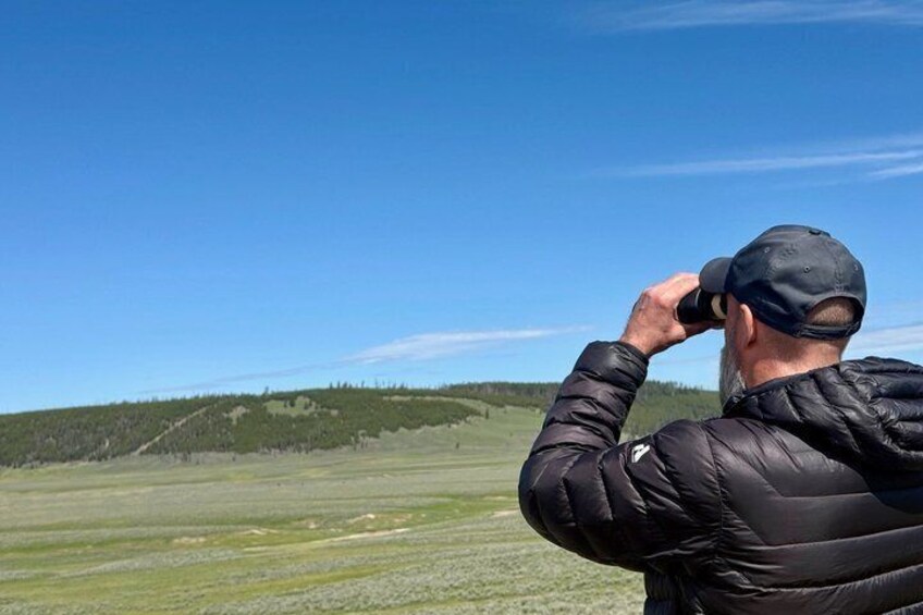 A person in a black jacket and cap looking through binoculars at a landscape with a clear sky and distant trees. 4