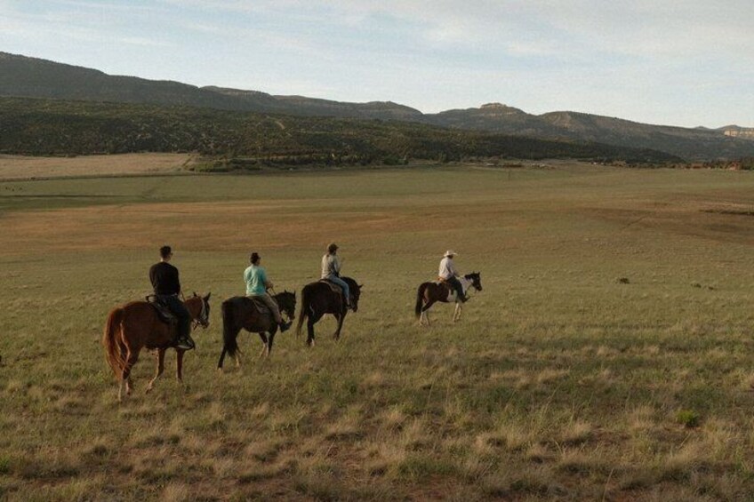 A group of people horseback riding across a grassy field with hills in the background. 3