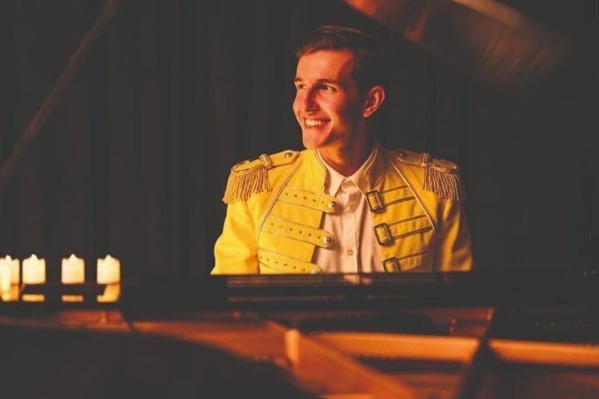 A person in a yellow military-style uniform with epaulettes, seated at a piano in a dimly lit room with candles. 1