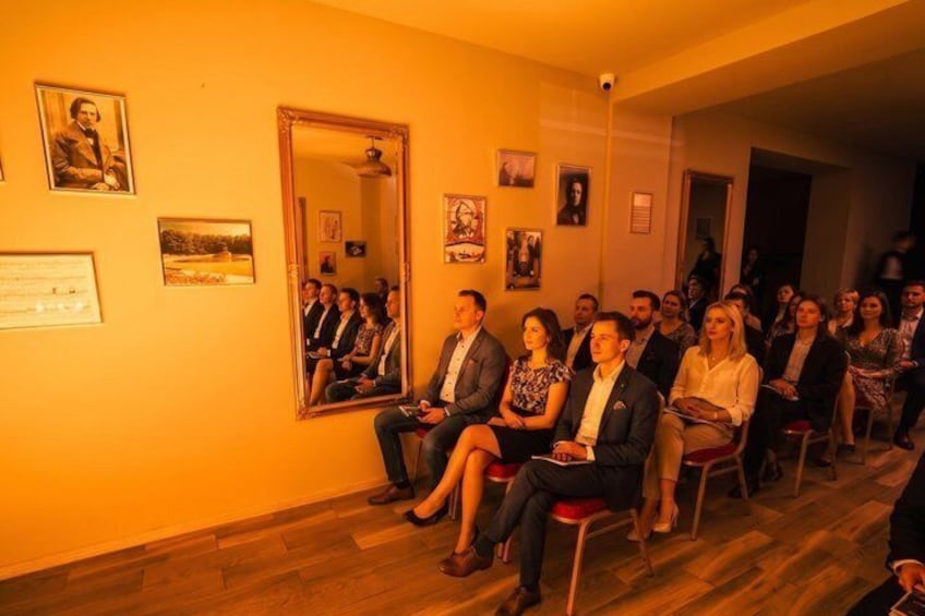 A group of people seated in chairs in a room with wooden floors and walls adorned with framed pictures. 4
