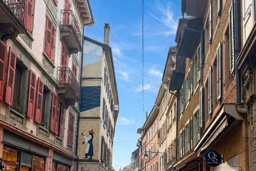 A narrow street with buildings on either side, some with balconies and shutters. 3