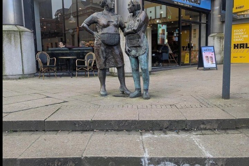 A bronze statue of two figures on a sidewalk, with a coffee shop and a signboard in the background. 2