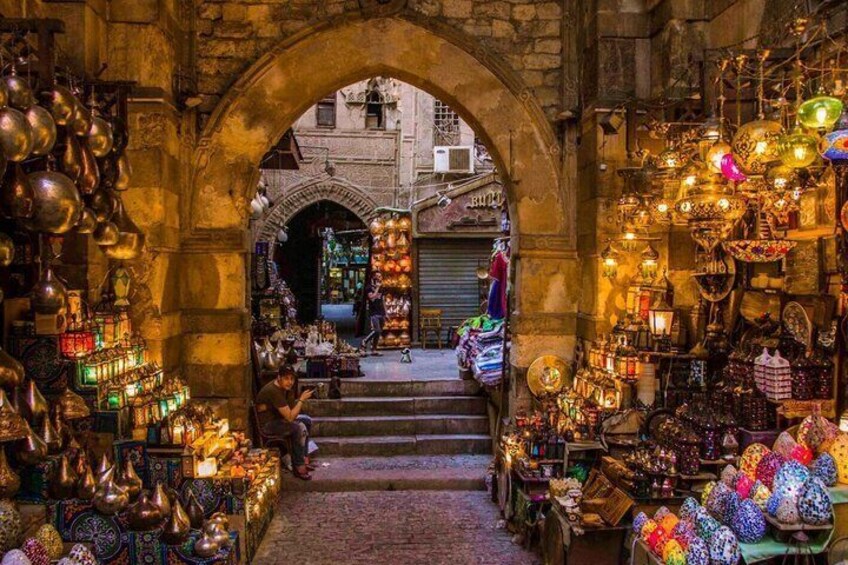 An indoor market with a stone archway, various stalls selling colorful items, and a person sitting on the steps. 2