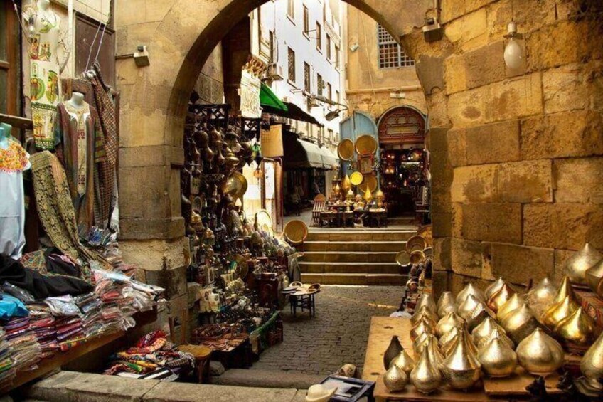 A narrow street with shops on either side, displaying various items like textiles and metal objects. 3