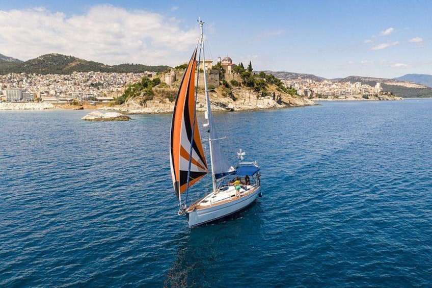 A sailboat with a striped sail on a calm sea, a coastal town in the background, and a hilly landscape. 1