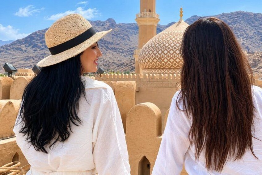 Two people facing a mosque with a dome and minaret, set against a mountainous backdrop. 1