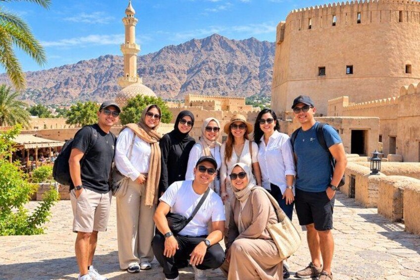 A group of people posing in front of a historical building with a minaret, palm trees, and mountains in the background. 3
