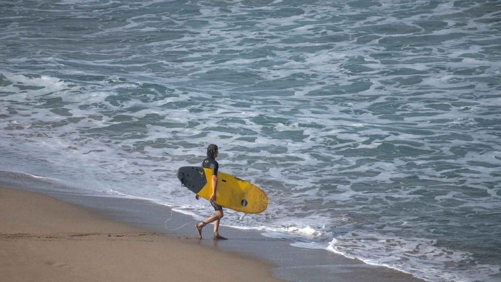 A person walking along a beach with waves crashing onto the shore, carrying a surfboard. 2