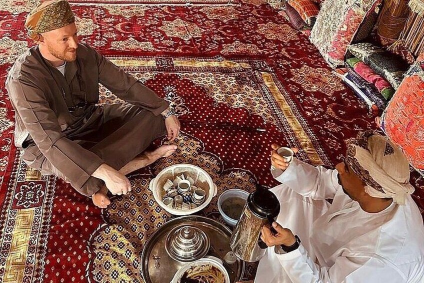 Two men sitting on a patterned carpet, one holding a cup. 3