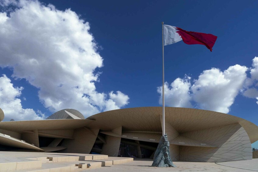 A modern architectural building with a unique curved design, a flagpole with a national flag, and a clear blue sky with clouds. 1