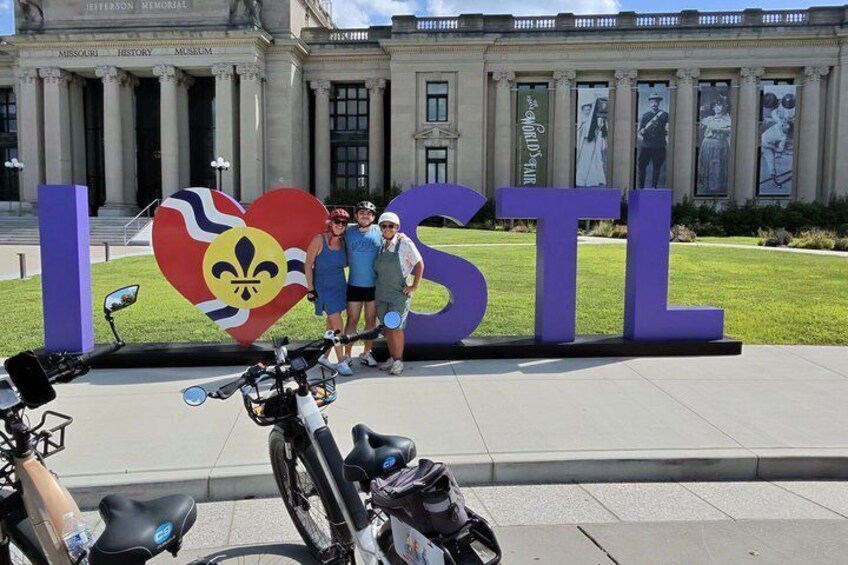 A large sign with 'I ♥ STL' and a fleur-de-lis symbol, in front of a building with columns. 3
