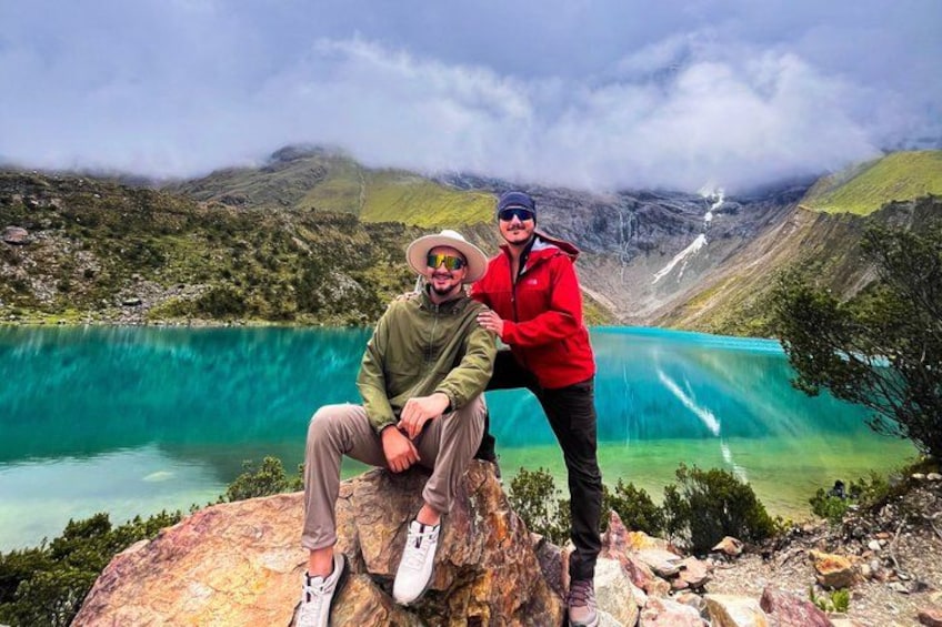 Two people sitting on a rock by a turquoise lake with a waterfall in the background. 1