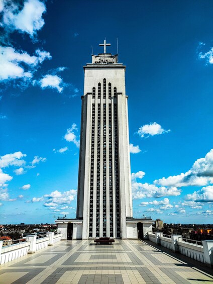 A tall, modern building with a cross on top, surrounded by a paved plaza and a railing. 3
