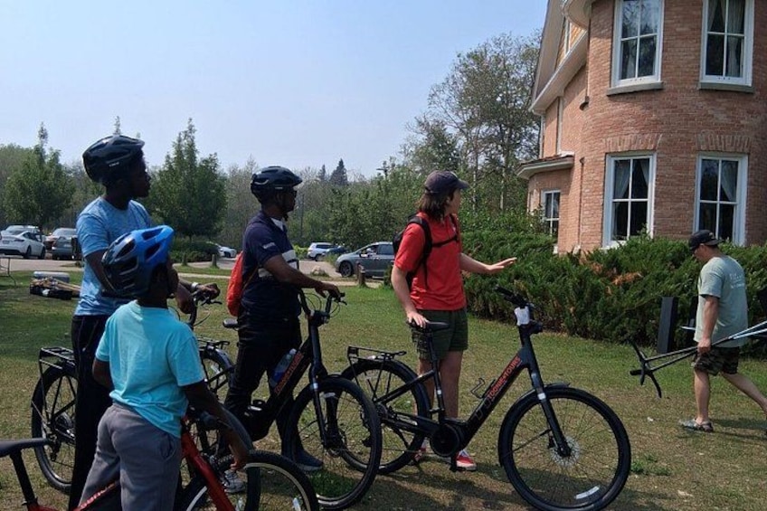 A group of people with bicycles in a grassy area, a brick building, and parked cars in the background. 2
