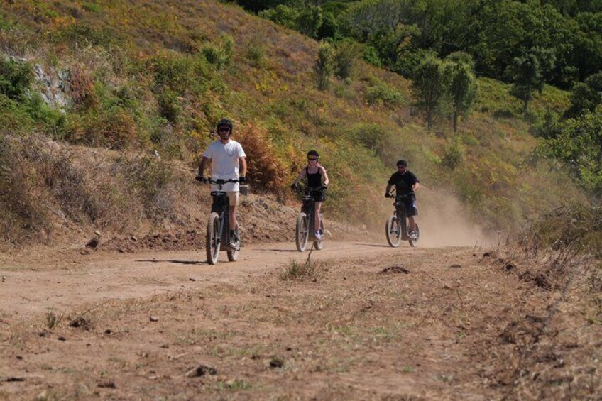 Three cyclists on a dirt path surrounded by dry vegetation and hills. 1