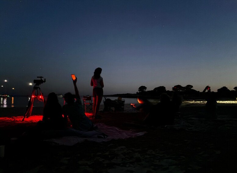 A beach at dusk with people gathered around a campfire, a telescope, and a boat in the background. 3