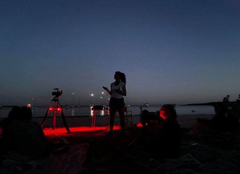 A person standing on a beach at dusk with a telescope and tripod, and several boats in the background. 1
