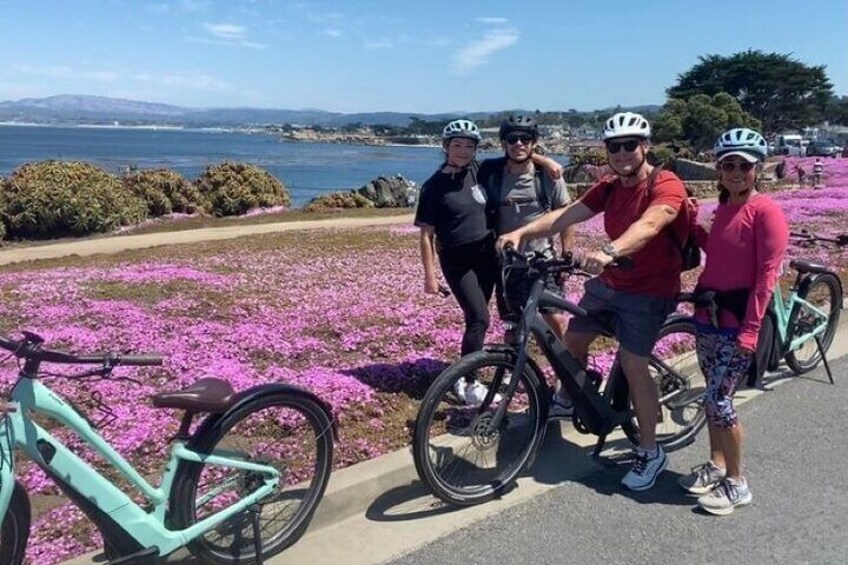 Un grupo de ciclistas en un sendero pavimentado con flores rosadas, una bicicleta turquesa y un pintoresco paisaje costero. 3