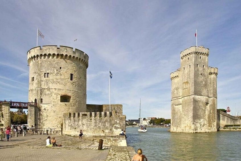 A historic stone fortress with two prominent towers, a paved walkway, and a body of water with boats. 2