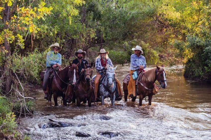 Four people riding horses across a shallow river surrounded by dense greenery. 1