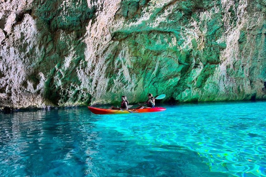 Two people kayaking in clear turquoise water with a rocky cliff in the background. 1