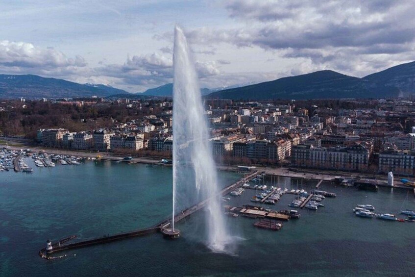 A cityscape with a prominent water fountain in the foreground, a marina with boats, and mountains in the background. 5