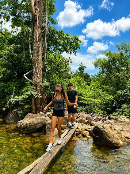Two people walking on a wooden bridge over a stream in a forested area with large rocks and trees. 1