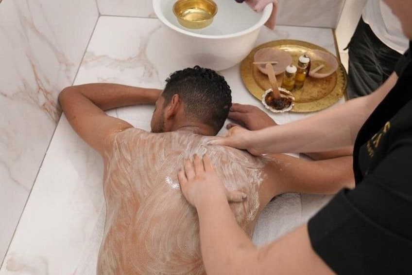 A person receiving a back massage in a marble-floored room with a basin and bottles on a tray. 1
