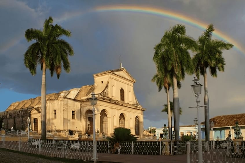 A historic building with a large arched doorway, palm trees, and a rainbow in the sky. 1